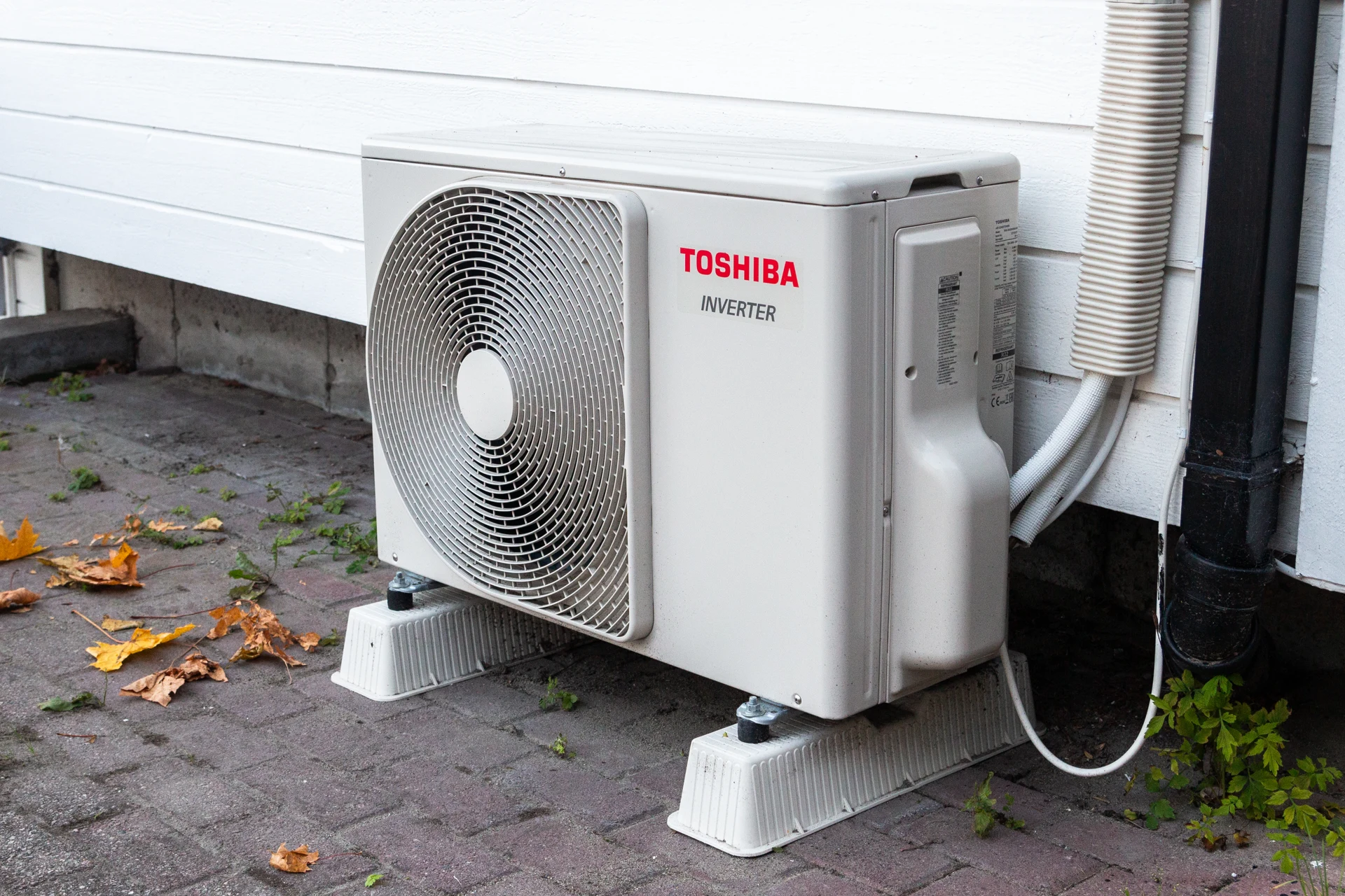 An outdoor residential heat pump unit raised on support feet beside white house siding.